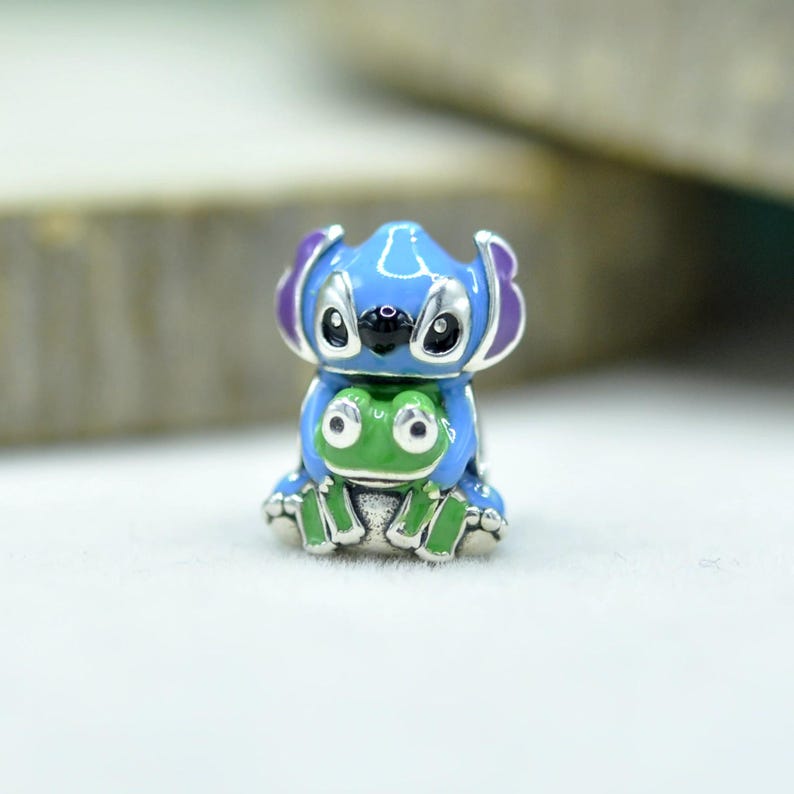 May include: A metal charm bead featuring a blue character with large ears and purple accents, holding a small green frog. The design includes black eyes and nose details, with silver-toned metal outlines defining the shapes of both figures.