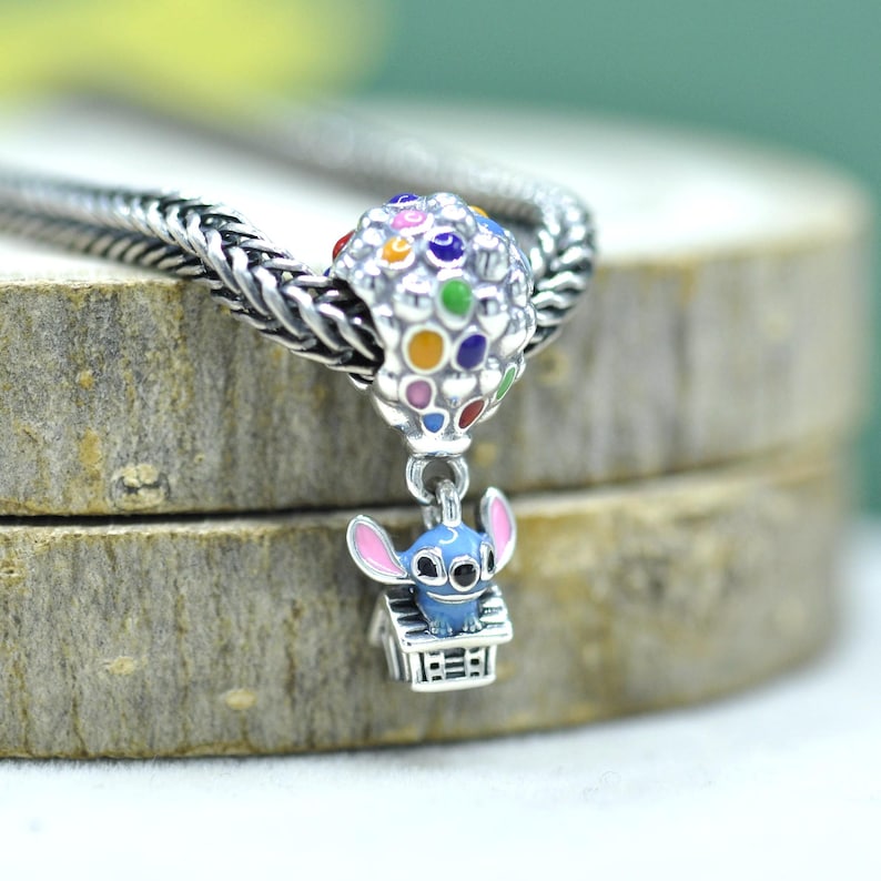 May include: A silver charm bead featuring a blue character with pink ears sitting on a small house, suspended from a larger silver bead decorated with colorful enamel dots. The charm is threaded onto a silver chain with a braided metal texture.