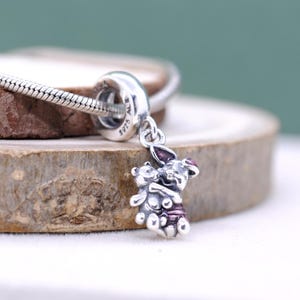Puede incluir: Un charm de plata con un oso y un conejo abrazándose. El conejo tiene detalles morados en las orejas y la ropa. El charm está unido a una pulsera de plata. El fondo es una superficie de madera.