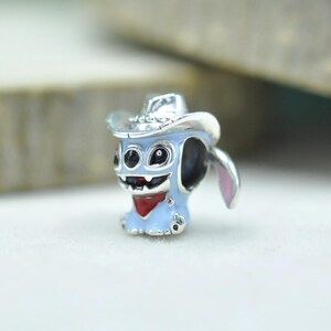 May include: A silver-toned metal charm bead shaped like a light blue character wearing a silver cowboy hat. The character features large black eyes, a wide grin with visible teeth, and a red bandana around its neck, with a pink inner ear detail.