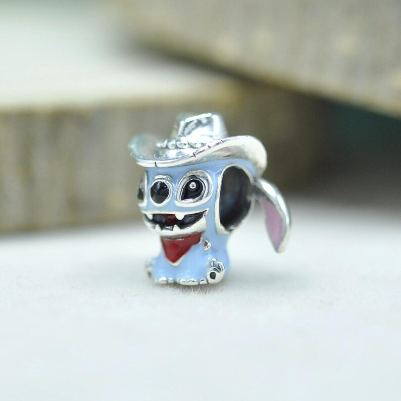 May include: A silver-toned metal charm bead shaped like a light blue character wearing a silver cowboy hat. The character features large black eyes, a wide grin with visible teeth, and a red bandana around its neck, with a pink inner ear detail.