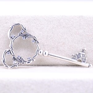 May include: A silver-toned key pendant with a decorative head featuring a floral design and embedded clear crystals. The key's handle has a castle design. The key is displayed on a light-colored surface.