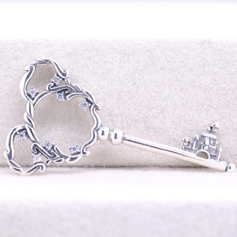 May include: A silver-toned key pendant with a decorative head featuring a floral design and embedded clear crystals. The key's handle has a castle design. The key is displayed on a light-colored surface.