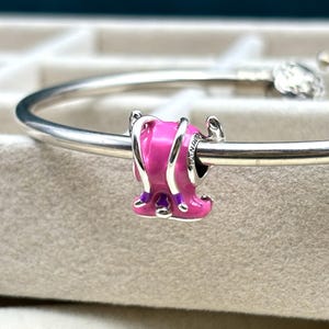 May include: A silver charm bracelet with a pink and purple enamel charm shaped like a shoe. The charm has a silver band around it and is attached to the bracelet.