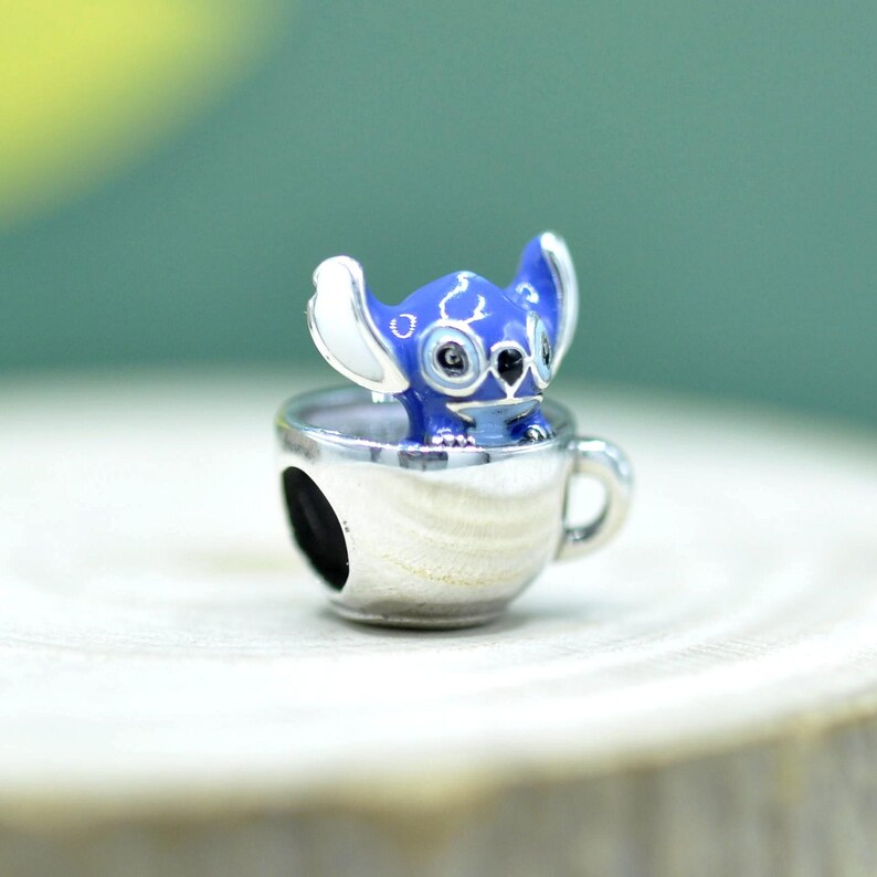 May include: A silver-toned metal charm featuring a blue and white enamel character with large ears sitting inside a small cup. The charm has a smooth, reflective finish and a circular opening on the side, designed to be threaded onto a bracelet or chain.
