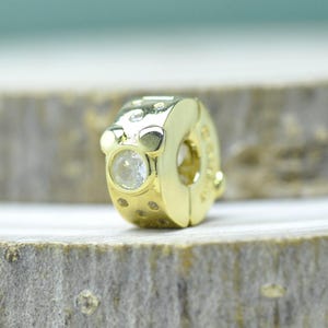 Puede incluir: Un charm de color dorado con una gema redonda y transparente engastada en la parte delantera. El charm tiene una forma redondeada con peque&ntilde;as hendiduras decorativas y una gran abertura lateral. El charm est&aacute; dise&ntilde;ado para sujetarse a una pulsera o collar.