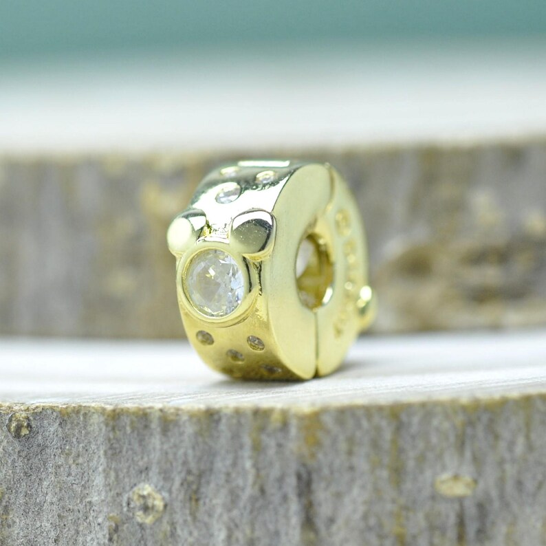 Puede incluir: Un charm de color dorado con una gema redonda y transparente engastada en la parte delantera. El charm tiene una forma redondeada con peque&ntilde;as hendiduras decorativas y una gran abertura lateral. El charm est&aacute; dise&ntilde;ado para sujetarse a una pulsera o collar.