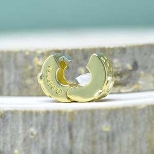 Puede incluir: Un cierre de joyer&iacute;a dorado, abierto y listo para ser conectado. El cierre tiene forma redondeada con una superficie texturizada y un peque&ntilde;o inserto de pl&aacute;stico blanco. La palabra "Disney" est&aacute; grabada en el cierre.