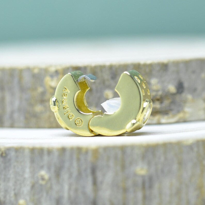 Puede incluir: Un cierre de joyer&iacute;a dorado, abierto y listo para ser conectado. El cierre tiene forma redondeada con una superficie texturizada y un peque&ntilde;o inserto de pl&aacute;stico blanco. La palabra "Disney" est&aacute; grabada en el cierre.