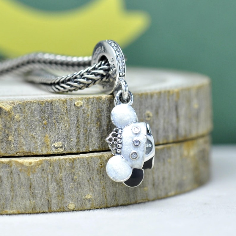 May include: A silver charm with a Mickey Mouse design. The charm features a white body, black ears, and a black and silver outfit. The charm is attached to a silver chain.