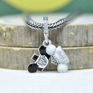 May include: A silver charm pendant featuring a black and white cartoon character couple, one in a tuxedo and the other in a wedding dress. The charm is suspended from a silver chain and has a decorative bail with clear stones.