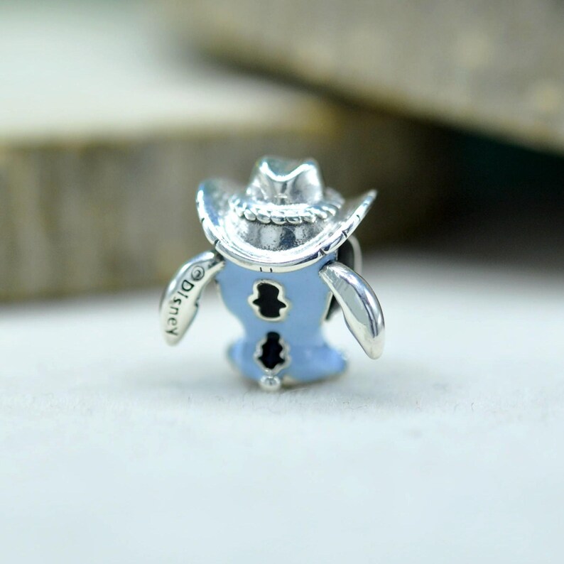 May include: A silver charm featuring a light blue enameled shirt with a cowboy hat on top. The piece includes a rope-textured brim on the hat, black cutout details on the shirt, and the word Disney engraved on the side of the silver sleeve.