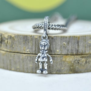 Op de afbeelding: Een zilveren bedel met een gedetailleerde figuur met hoed, jas en laarzen. De bedel hangt aan een gevlochten ring met kleine, sprankelende accenten. Geschikt voor een armband of ketting.