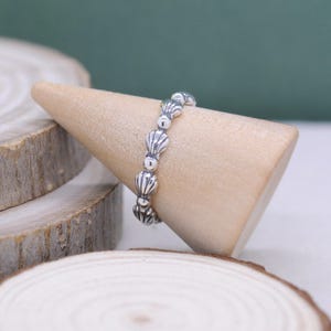 May include: A silver ring featuring a repeating seashell design. The ring is displayed on a wooden cone, showcasing its intricate details. The ring's design is a series of small, detailed seashells, creating a coastal or nautical theme.