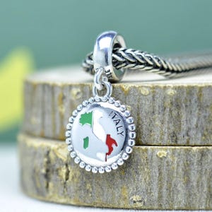 May include: A silver charm with a round pendant featuring the map of Italy in green, white, and red. The word "ITALY" is written around the edge of the pendant. The charm is attached to a braided silver chain.