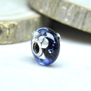 May include: A blue and white glass bead with silver accents. The bead has a dark blue background with white swirls and silver flower-shaped embellishments. The bead is a charm for a bracelet or necklace.