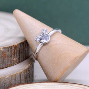 Può includere: Un anello d'argento con un design ispirato a Mickey Mouse. L'anello presenta una grande gemma rotonda e trasparente come faccia, con due gemme più piccole sopra e ai lati, che formano le orecchie. L'anello è esposto su un cono di legno.