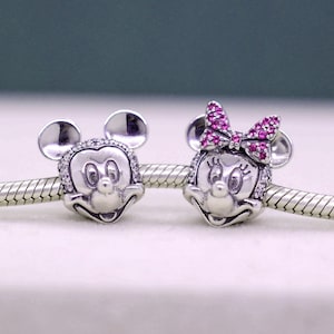 May include: Two silver charms featuring Mickey and Minnie Mouse. Mickey has a smiling face with a clear crystal border. Minnie has a pink crystal bow and a clear crystal border. Both charms have round ears.