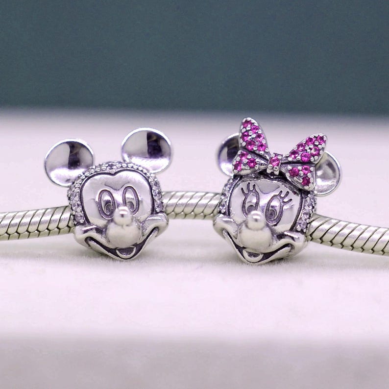 May include: Two silver charms featuring Mickey and Minnie Mouse. Mickey has a smiling face with a clear crystal border. Minnie has a pink crystal bow and a clear crystal border. Both charms have round ears.