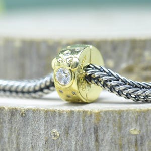 Puede incluir: Un charm de color dorado con una gema redonda y transparente y piedras transparentes m&aacute;s peque&ntilde;as. El charm est&aacute; unido a una cadena trenzada de color plateado. El charm tiene una superficie texturizada y es probablemente un componente de joyer&iacute;a.