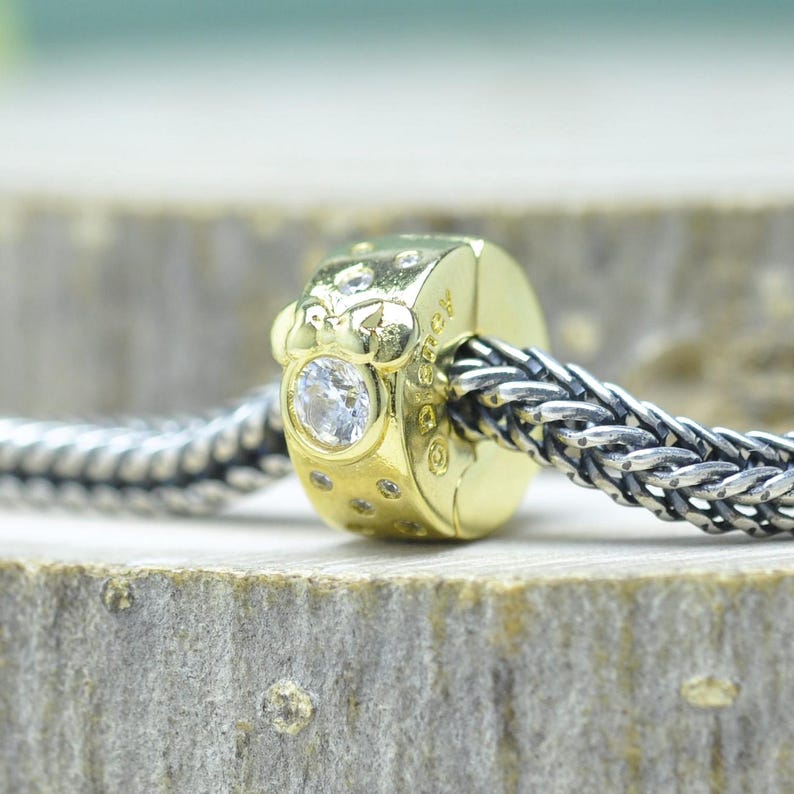 Puede incluir: Un charm de color dorado con una gema redonda y transparente y piedras transparentes m&aacute;s peque&ntilde;as. El charm est&aacute; unido a una cadena trenzada de color plateado. El charm tiene una superficie texturizada y es probablemente un componente de joyer&iacute;a.