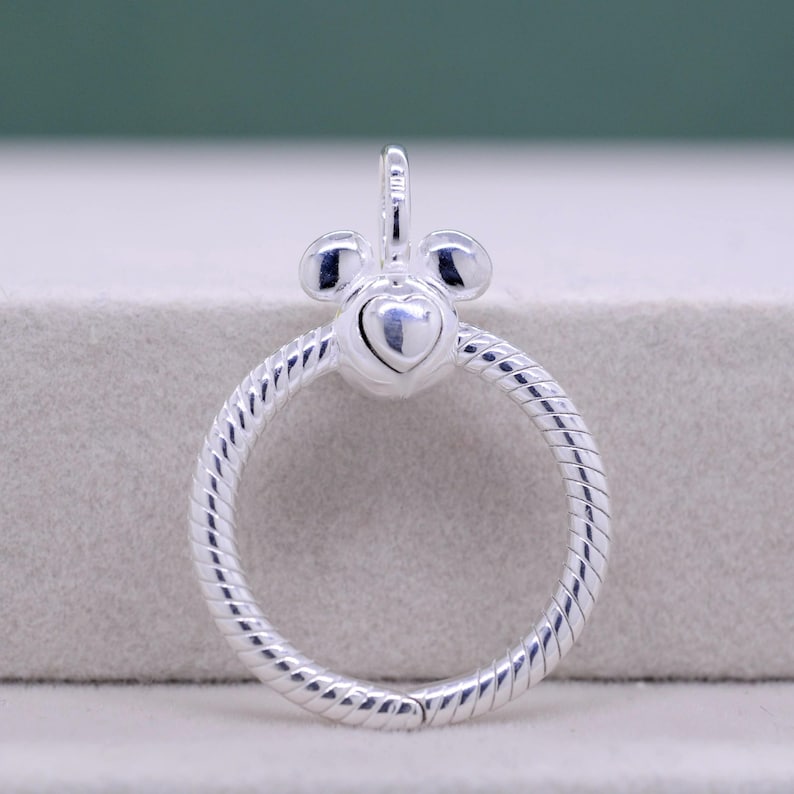 Pu&ograve; includere: Ciondolo color argento con un design a forma di cuore al centro e un anello circolare strutturato. Il ciondolo ha un piccolo anello nella parte superiore per appenderlo. Il design include due piccoli cerchi sopra il cuore.