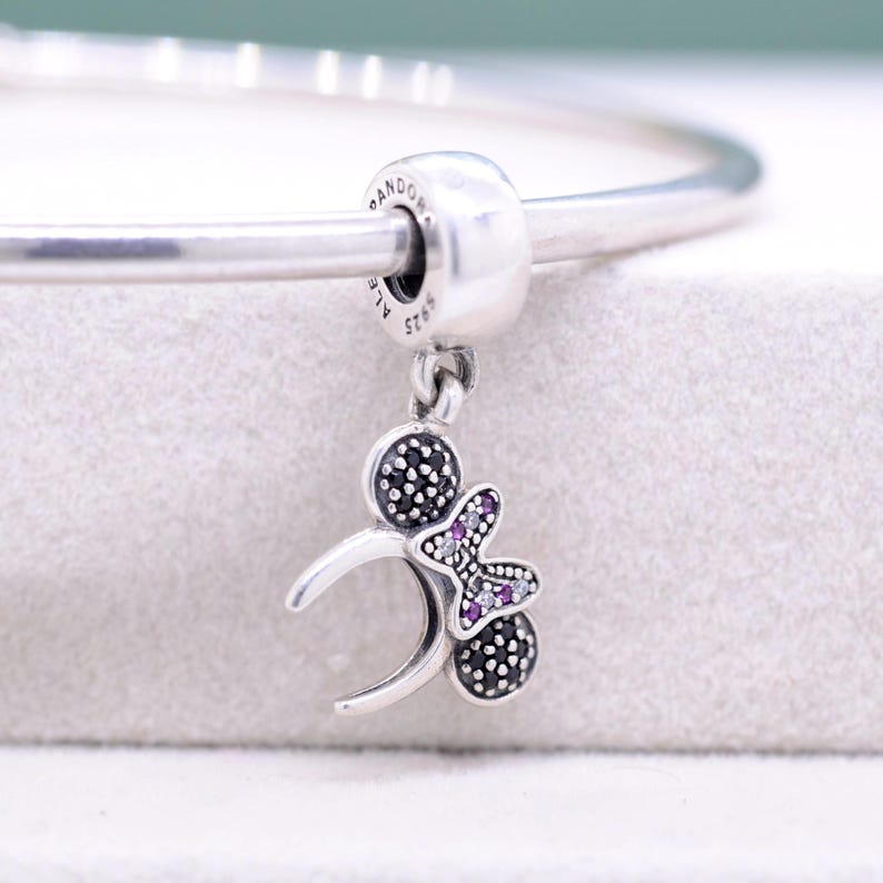 May include: A silver charm with a Mickey Mouse-inspired design. The charm features two black, studded circles and a butterfly with purple gems. The charm is attached to a silver bracelet. The charm is approximately 1 inch in length.