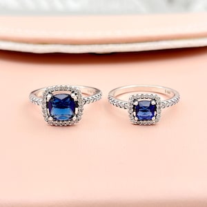 May include: Two silver rings, each featuring a square, deep blue gemstone at the centre. The stones are encircled by smaller, clear gems. The bands have a textured pattern. The rings are set against a pink backdrop.