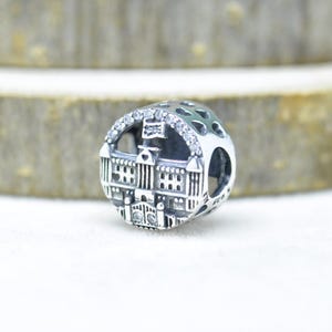 May include: Silver charm with a detailed architectural design. The charm features a building facade with windows and decorative elements, accented with clear crystals. The charm is round with a hollowed-out design on the side.