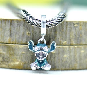 May include: A blue and silver charm featuring a cartoon character with large ears and a wide grin. The charm is suspended from a silver braided chain. The charm has white accents on its feet.