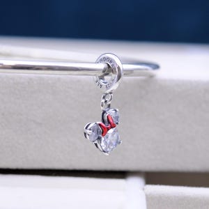 May include: Silver charm with a red bow and clear crystal accents, shaped like Minnie Mouse. The charm is attached to a silver bracelet with the word "Pandora" engraved on the clasp.