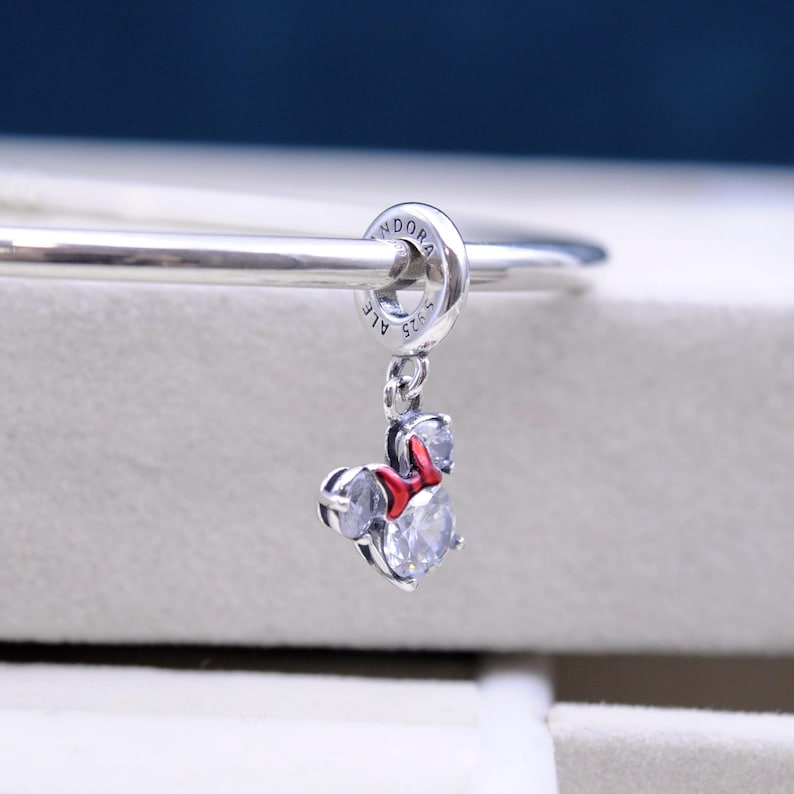 May include: Silver charm with a red bow and clear crystal accents, shaped like Minnie Mouse. The charm is attached to a silver bracelet with the word "Pandora" engraved on the clasp.