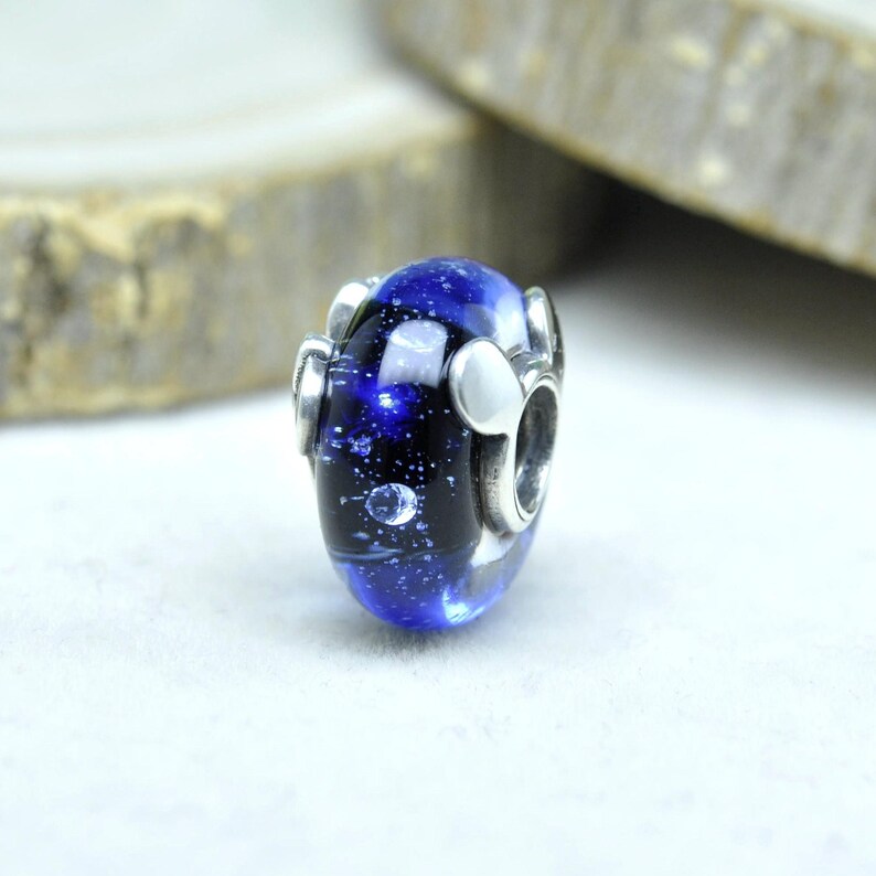 May include: A blue and black glass bead with a galaxy design. The bead has silver-colored metal accents and a hole for stringing. The bead is oval-shaped and has a shiny, reflective surface.