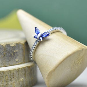 Peut inclure: Une bague en argent ornée d'un motif papillon avec des pierres bleues. Le papillon a un corps argenté et des ailes bleues. L'anneau est orné de petites pierres transparentes. La bague est présentée sur un support en bois.