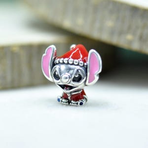 Puede incluir: Un charm de plata con la forma del personaje Stitch, que lleva un gorro de Pap&aacute; Noel rojo y un abrigo rojo. El personaje tiene grandes orejas con interior rosa, ojos negros y una amplia sonrisa. Los detalles en esmalte rojo son vibrantes.