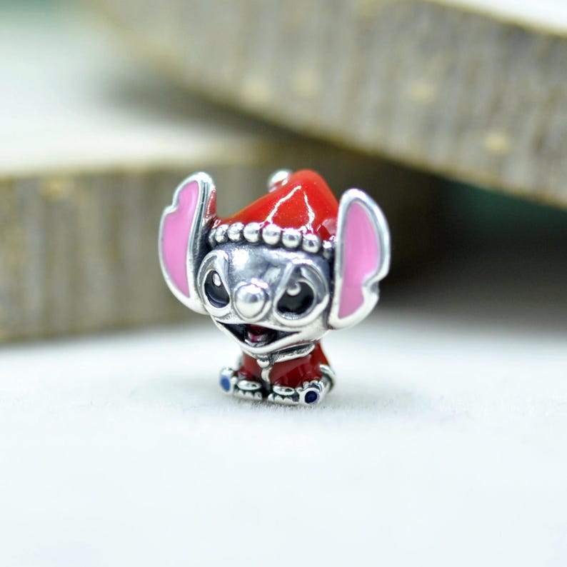Puede incluir: Un charm de plata con la forma del personaje Stitch, que lleva un gorro de Pap&aacute; Noel rojo y un abrigo rojo. El personaje tiene grandes orejas con interior rosa, ojos negros y una amplia sonrisa. Los detalles en esmalte rojo son vibrantes.