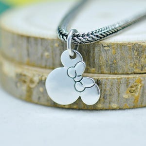 Peut inclure: Un collier pendentif argent&eacute; avec un motif Minnie Mouse. Le pendentif est une silhouette de la t&ecirc;te de Minnie avec un n&oelig;ud. La cha&icirc;ne du collier est en argent tiss&eacute; et textur&eacute;. Le pendentif est suspendu sur un fond en bois.