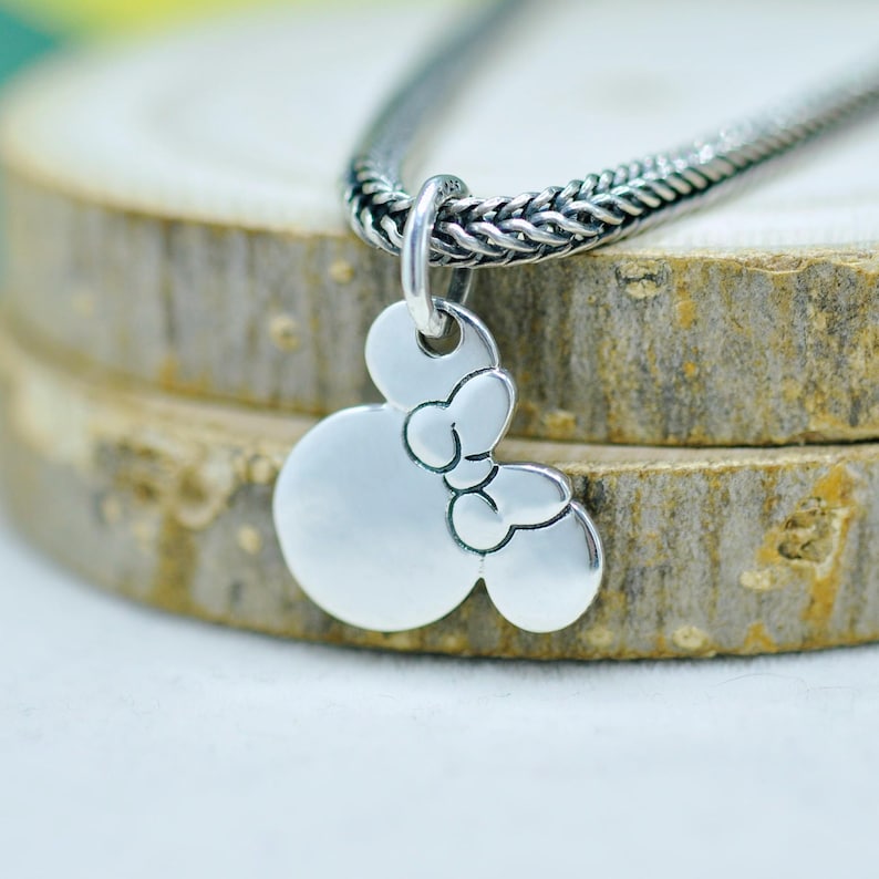 Peut inclure: Un collier pendentif argent&eacute; avec un motif Minnie Mouse. Le pendentif est une silhouette de la t&ecirc;te de Minnie avec un n&oelig;ud. La cha&icirc;ne du collier est en argent tiss&eacute; et textur&eacute;. Le pendentif est suspendu sur un fond en bois.