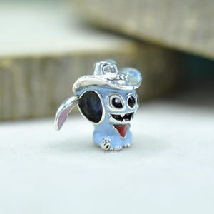 May include: A silver-toned metal charm bead shaped like a light blue character wearing a silver cowboy hat. The character features large black eyes, a wide grin, a red neckerchief, and a small pink-lined ear extending from the side of the head.