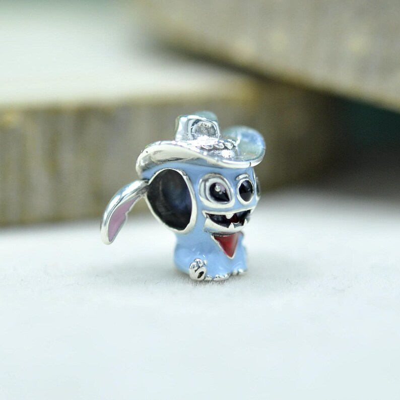 May include: A silver-toned metal charm bead shaped like a light blue character wearing a silver cowboy hat. The character features large black eyes, a wide grin, a red neckerchief, and a small pink-lined ear extending from the side of the head.