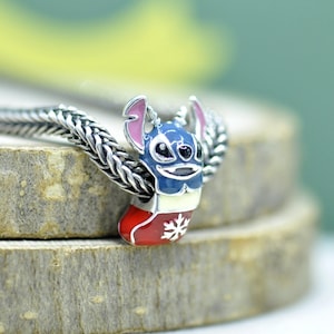 Puede incluir: Un abalorio de plata que muestra un personaje azul con orejas grandes y detalles rosas, saliendo de un calcet&iacute;n navide&ntilde;o rojo decorado con un copo de nieve blanco. El abalorio est&aacute; ensartado en una cadena de plata con textura trenzada.