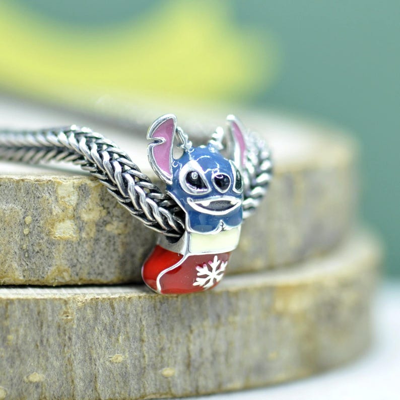 Puede incluir: Un abalorio de plata que muestra un personaje azul con orejas grandes y detalles rosas, saliendo de un calcet&iacute;n navide&ntilde;o rojo decorado con un copo de nieve blanco. El abalorio est&aacute; ensartado en una cadena de plata con textura trenzada.