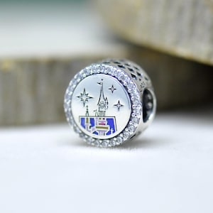 Può includere: Un charm in argento con un design dettagliato di un castello. Il charm presenta un design circolare centrale con un'illustrazione del castello in blu, viola e bianco, circondato da cristalli trasparenti. Il charm ha un motivo a stelle e diamanti sul lato.