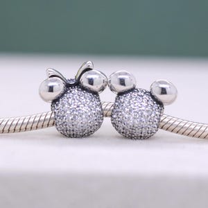 May include: Two silver-toned charm beads, one with a bow, shaped like cartoon characters. The charms are encrusted with small, sparkling crystals. They are displayed on a silver chain, perfect for a bracelet or necklace.
