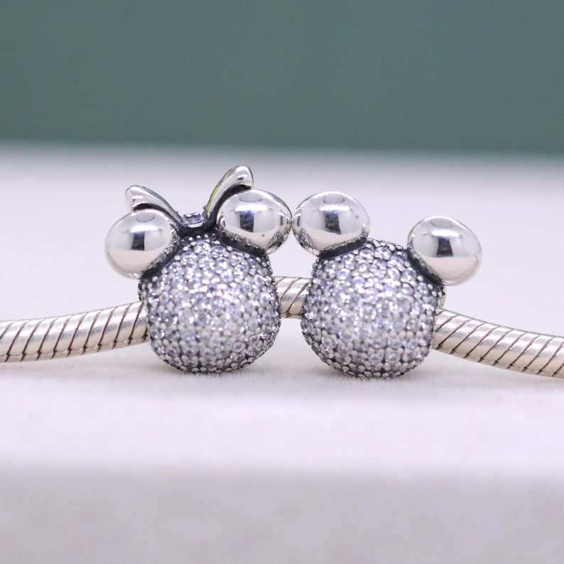 May include: Two silver-toned charm beads, one with a bow, shaped like cartoon characters. The charms are encrusted with small, sparkling crystals. They are displayed on a silver chain, perfect for a bracelet or necklace.