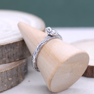 Könnte beinhalten: Ein silberner Ring mit einem Vogel auf einem Ast. Das Band des Rings ist strukturiert, um Baumrinde nachzuahmen, und der Vogel hat detaillierte Merkmale. Der Ring wird auf einem Holzkegel präsentiert.