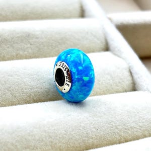 May include: A blue and green opal charm bead with a silver base. The bead has a silver charm with the inscription "S925 ALE" on it.