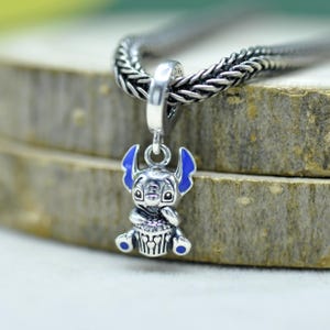 May include: A silver charm pendant featuring a cartoon character with blue ears and accents. The charm is suspended from a braided silver chain. The character is holding a cupcake. The charm is approximately 1 inch in height.
