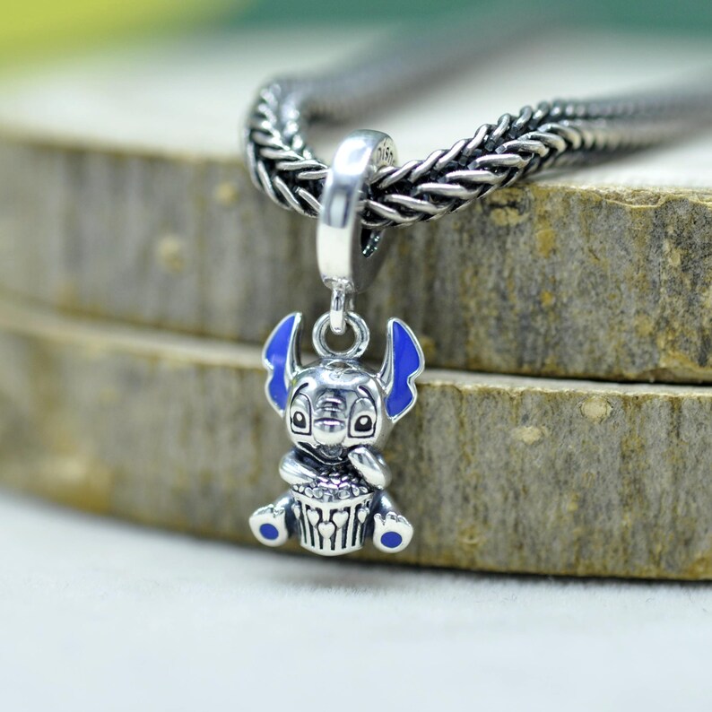 May include: A silver charm pendant featuring a cartoon character with blue ears and accents. The charm is suspended from a braided silver chain. The character is holding a cupcake. The charm is approximately 1 inch in height.