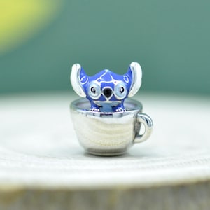 May include: A silver-toned metal charm featuring a blue enamel character with large ears and dark eyes, positioned inside a small silver-toned cup. The character has white accents on the ears and a smooth, polished finish on the cup exterior.